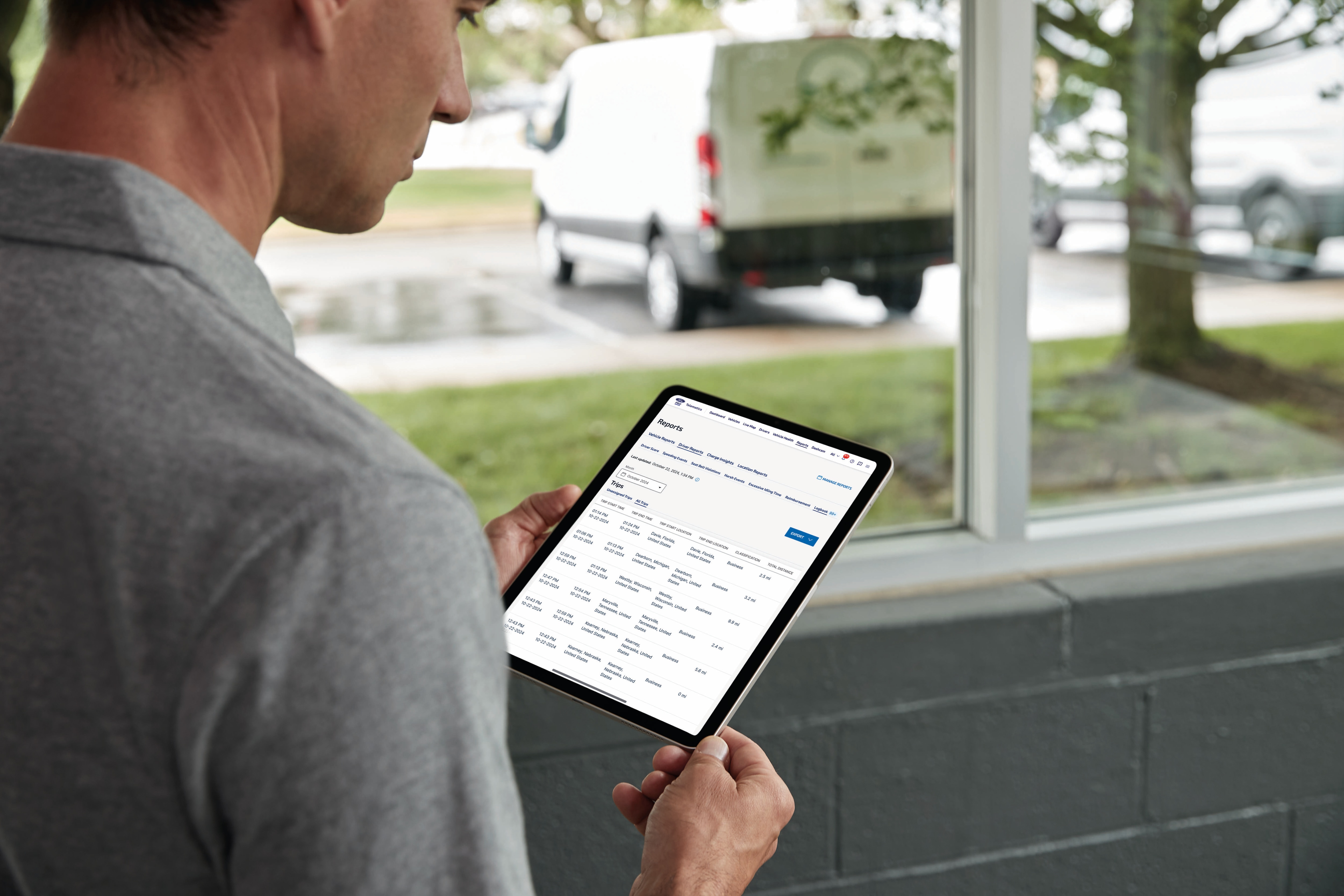 A man holds a tablet in front of a window. Outside the window, a van sits in the parking lot.